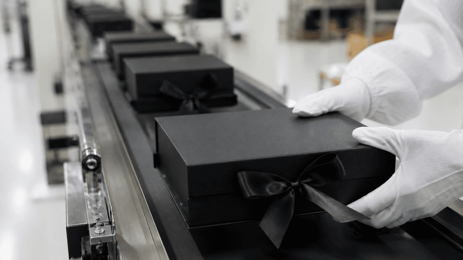 Photograph of a clean factory production line with rows of premium black cosmetic gift boxes moving along a conveyor in soft focus, with one box in the foreground in sharp focus being inspected by gloved hands, bright clean industrial lighting with neutral palette