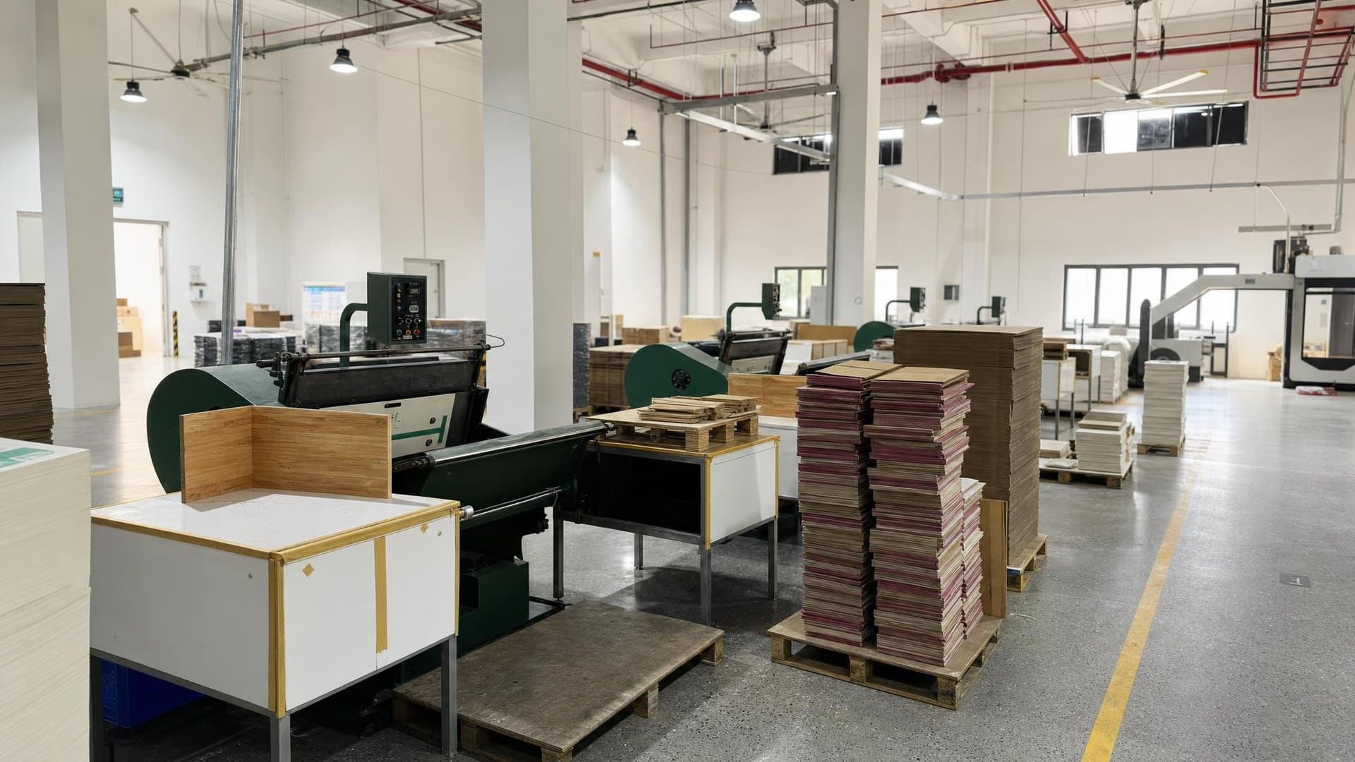 Printed carton sheets leaving a six-color offset press in an enclosed printing hall, with die-cut and folded cartons stacked beside the delivery end
